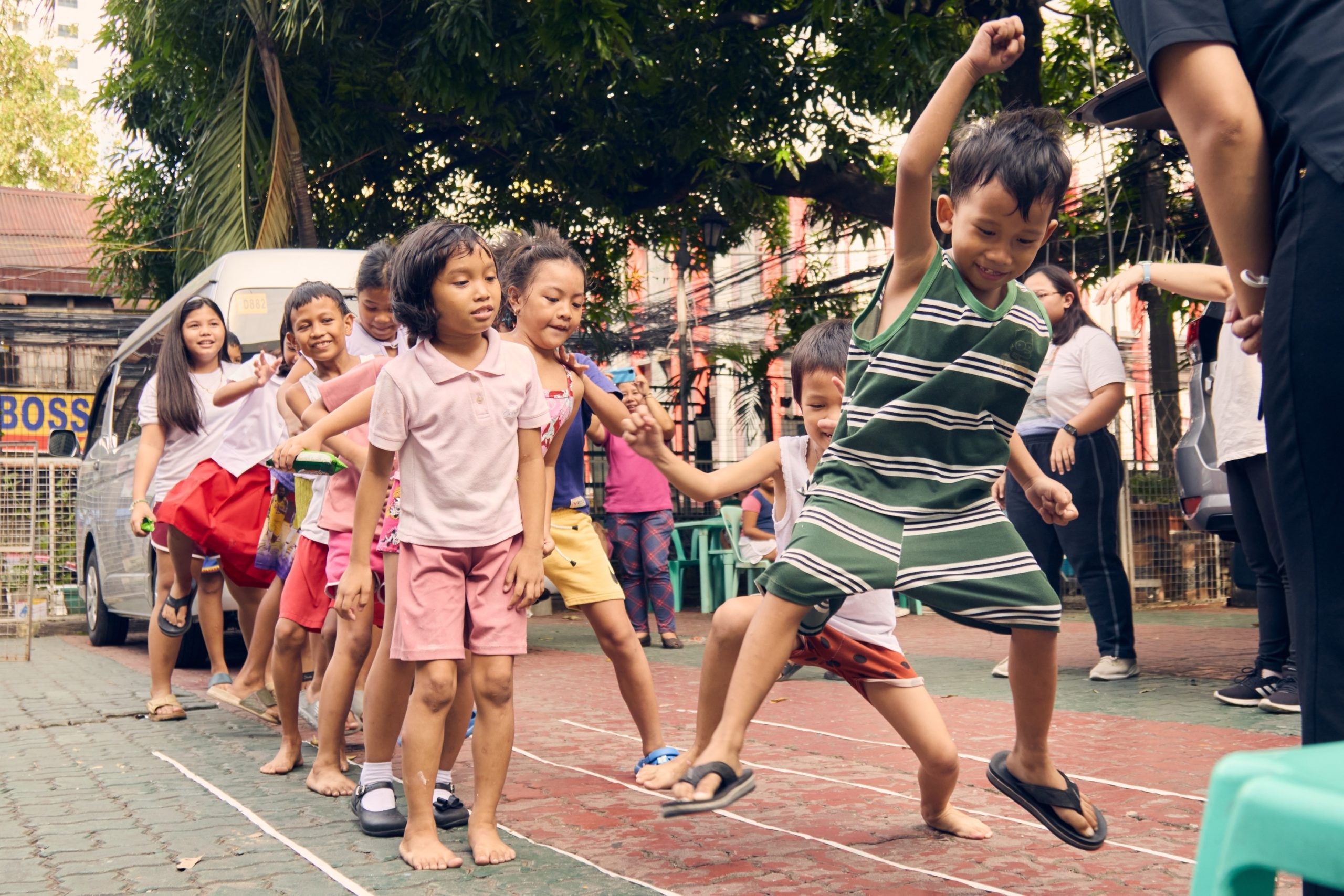 A group of boys and girls play an outdoor game, jumping over white lines on the ground.