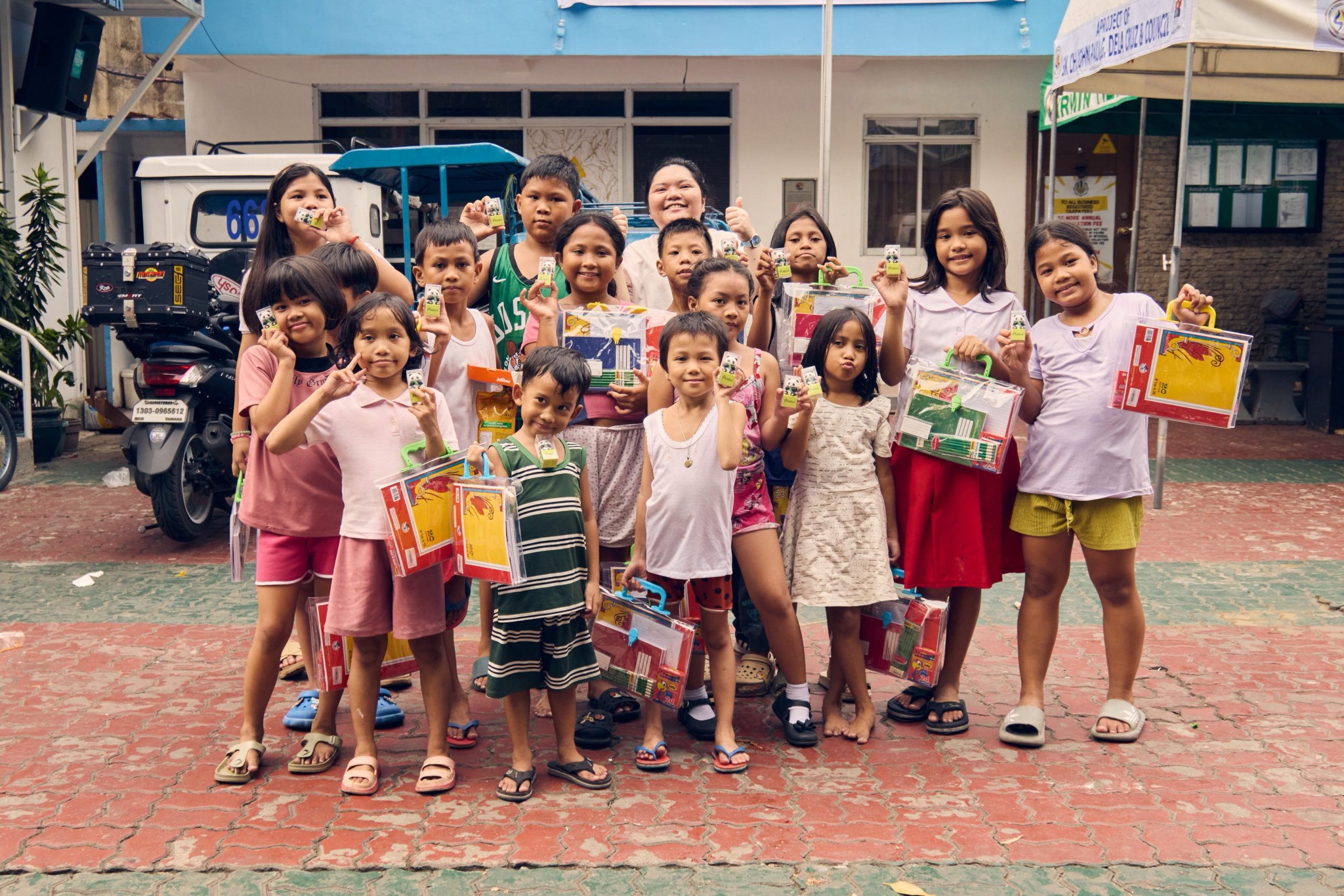 A group of children, along with an adult, are standing together outdoors, holding school supplies and small items.